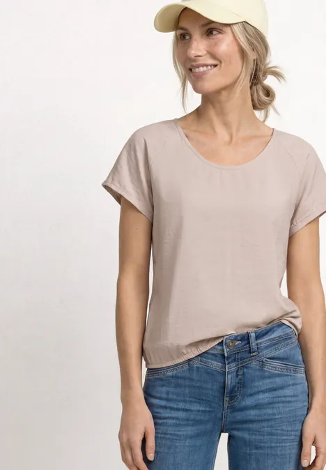Jonge vrouw met beige t-shirt en blauwe jeans tegen neutrale achtergrond.