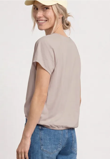 Een model in beige t-shirt en blauwe jeans tegen een witte achtergrond zijdelings gefotografeerd