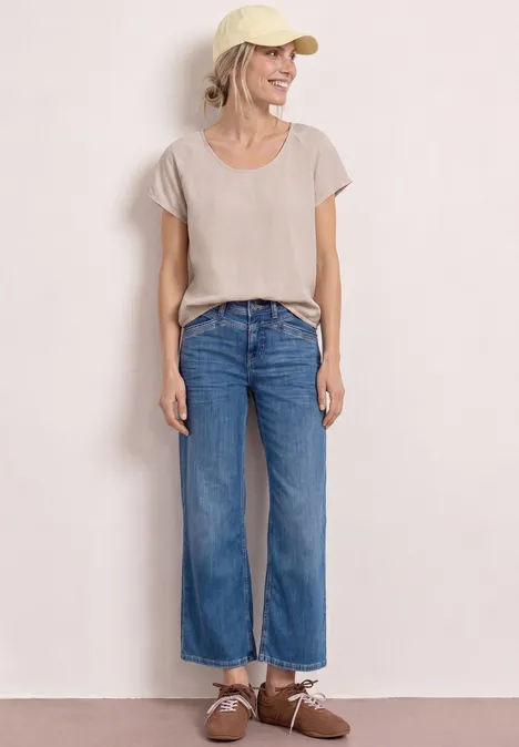 Een persoon staat voor een witte achtergrond, draagt een beige t-shirt, rechte blauwe jeans en bruine schoenen.