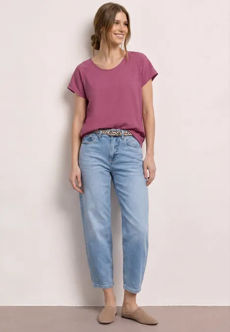 Een persoon draagt een paars t-shirt, lichtblauwe jeans met riem en bruine mules op lichte achtergrond.