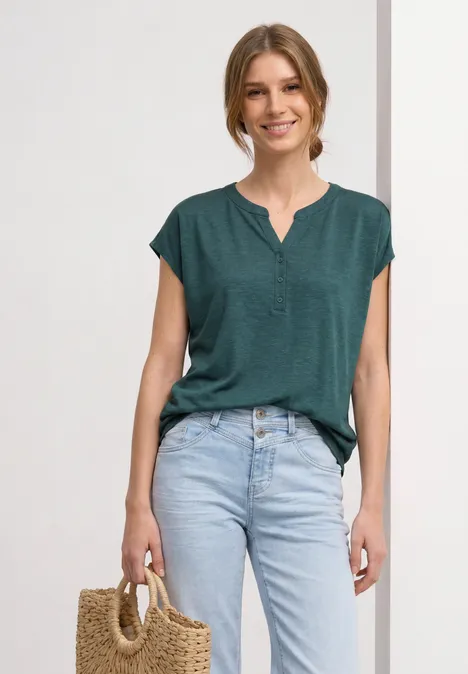 Dropped Shoulder Shirt mit Split Neck mediterra green Frau mit grüner Kurzarmbluse, hellblauen Jeans und Strohtasche vor neutralem Hintergrund.