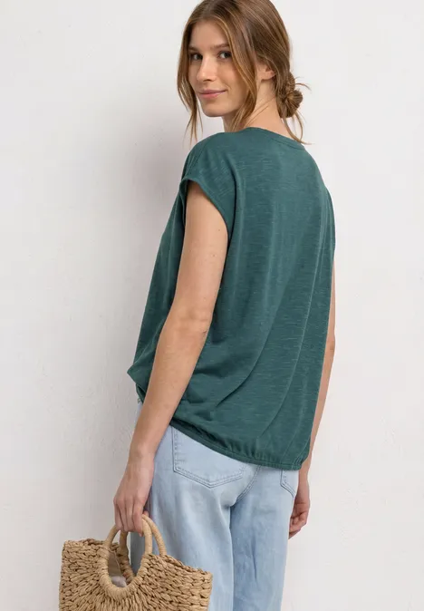 Dropped Shoulder Shirt mit Split Neck mediterra green Frau in lockerem tealgrünen Shirt und Jeans, seitlich stehend mit Strohtasche vor hellem Hintergrund