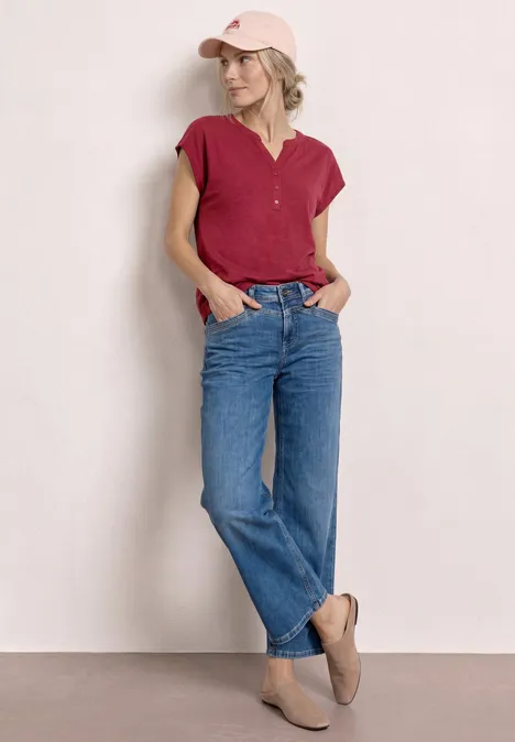 Dropped Shoulder Shirt mit Split Neck truffle red Frau lehnt entspannt an der Wand in rotem Oberteil, geraden blauen Jeans und hellbraunen Slippern.