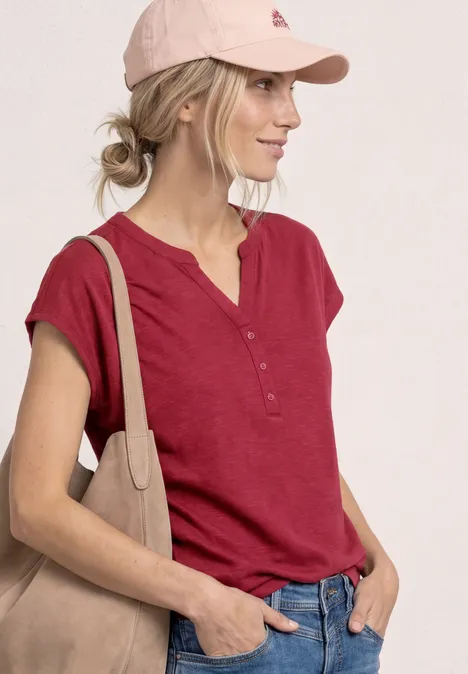 Dropped Shoulder Shirt mit Split Neck truffle red Frau mit rotem Kurzarmhemd, rosa Cap und beiger Schultertasche vor hellem Hintergrund.