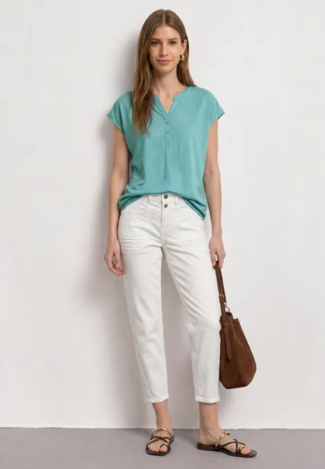 Dropped Shoulder Shirt mit Split Neck misty mint Frau steht vor weißer Wand, trägt türkisfarbenes Oberteil, weiße Hose, schwarze Sandalen und braune Tasche.