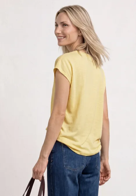 Een vrouw van achteren in een geel T-shirt en blauwe jeans, met een handtas in de hand.