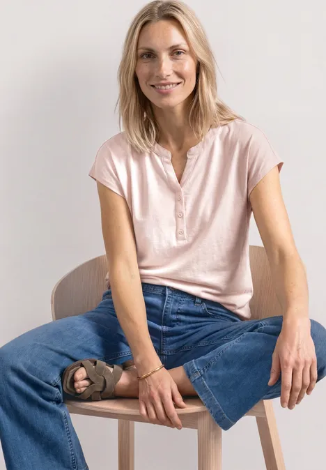 Dropped Shoulder Shirt mit Split Neck minimal rose Frau sitzt auf Stuhl in rosa Shirt und weiten Jeans, ein Bein angehoben.