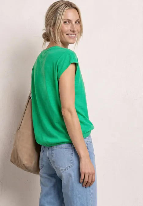 Dropped Shoulder Shirt mit Split Neck ever green Frau im Profil trägt grünes Shirt, blaue Jeans und eine beige Umhängetasche vor neutralem Hintergrund.