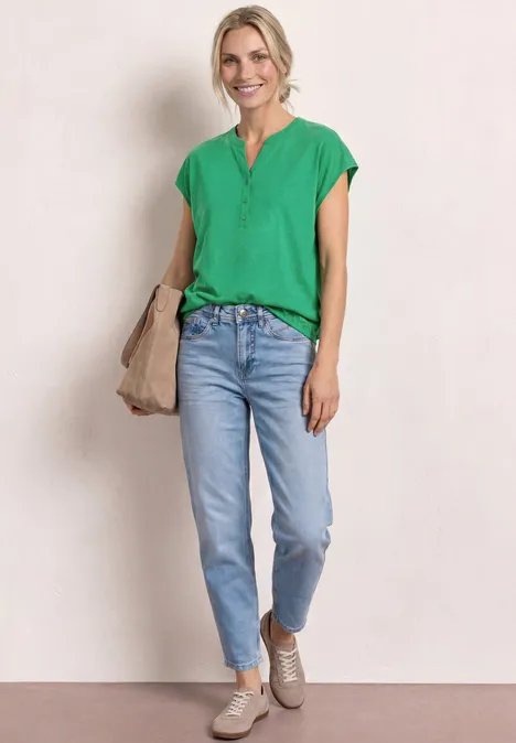 Dropped Shoulder Shirt mit Split Neck ever green Frau in grünem Oberteil und hellblauen Jeans, trägt beige Tasche und Schuhe vor hellem Hintergrund.