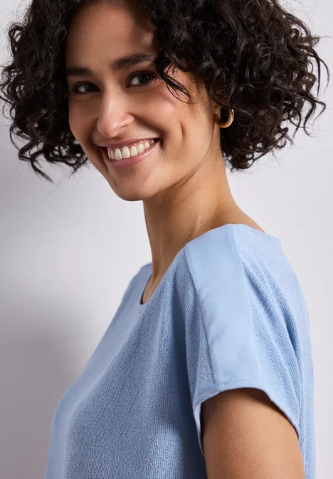 Eine Person mit lockigen Haaren trägt ein hellblaues, kurzärmliges Oberteil.