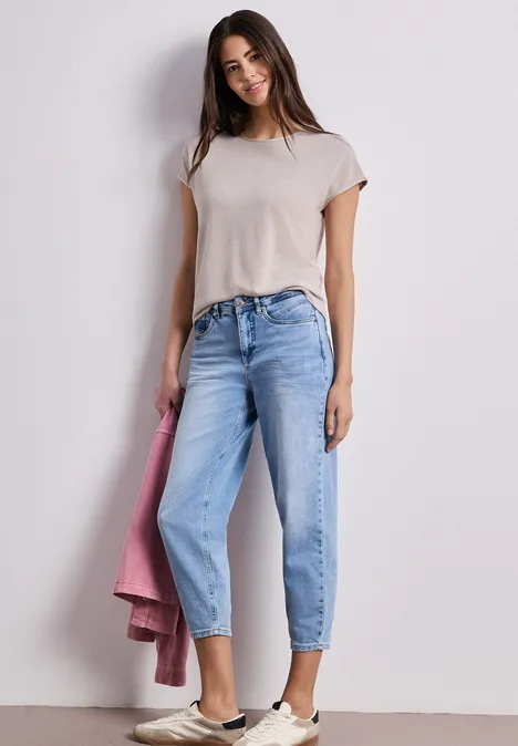 Junge Frau in weißem T-Shirt, locker sitzenden hellblauen Jeans und weißen Sneakern, hält rosa Jacke.