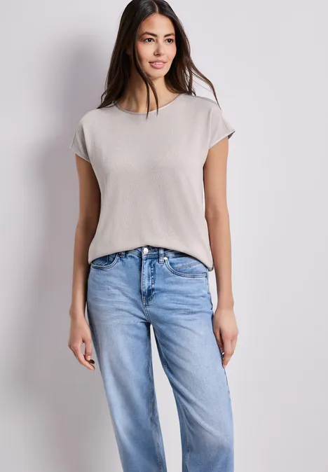 Eine Frau steht vor weißem Hintergrund und trägt ein schlichtes weißes T-Shirt und blaue Jeans.