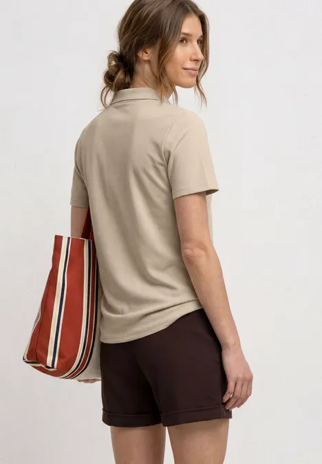 Eine Frau steht im Profil, trägt ein beiges Poloshirt, braune Shorts und eine rot-weiß gestreifte Tasche.