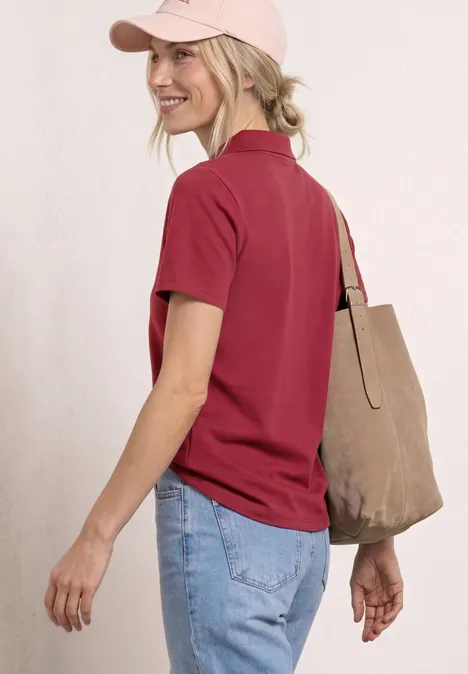 Eine Frau von hinten in rotem Poloshirt, blauer Jeans, rosa Baseballcap und beiger Stofftasche.