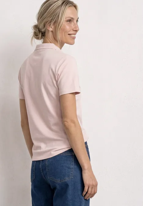 Vrouw van achteren in een roze T-shirt en blauwe jeans tegen een effen lichte achtergrond.