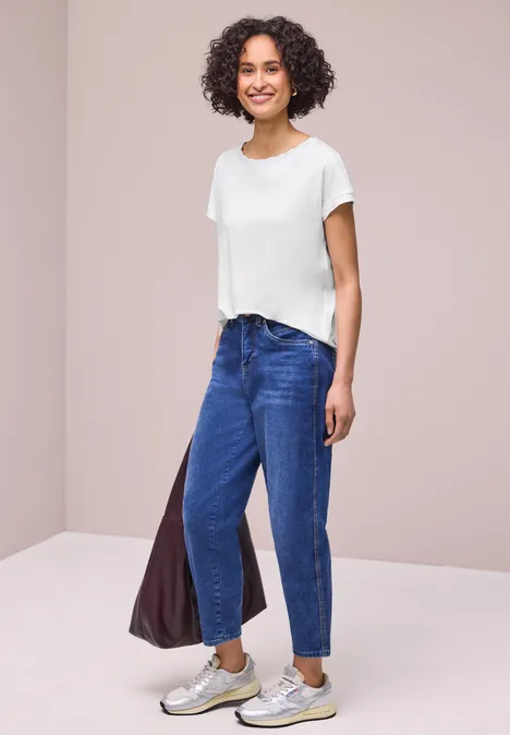 Eine Person trägt ein weißes T-Shirt, blaue Jeans, Sneaker und hält eine Tasche in neutralem Raum.