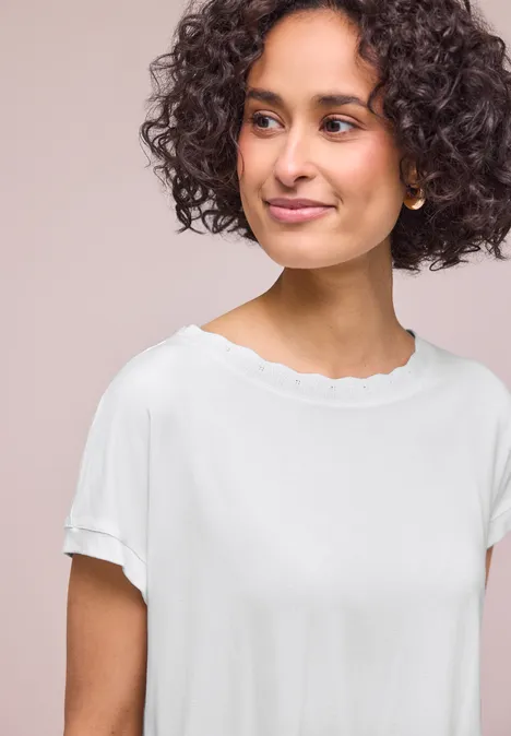 Person mit lockigem Haar trägt ein weißes T-Shirt mit wellenförmigem Halsausschnitt und umgekrempelten Ärmeln.