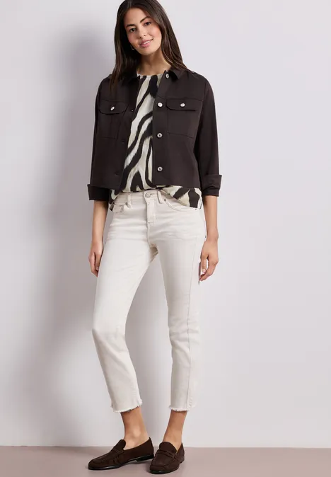 Dame in weißer Jeans, schwarzer Kurzjacke, gemusterter Bluse und braunen Loafers vor hellem Hintergrund.