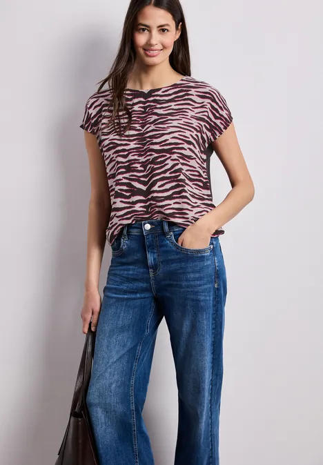 Frau trägt Zebra-Print-Shirt, blaue Jeans und schwarze Tasche vor hellem Hintergrund.