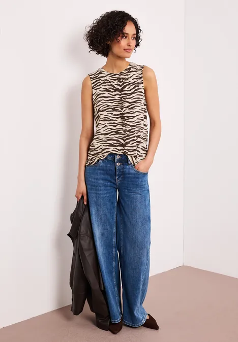 Vrouw in mouwloze zebraprint top, wijde jeans en zwarte jas voor een witte muur.