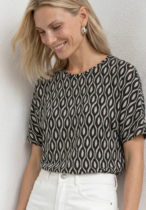 Kurzarm Shirt mit Rundhals und Print clay sand Frau trägt gemustertes, schwarz-beige Oberteil und weiße Hose.