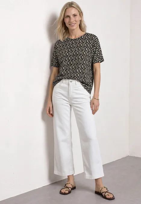 Kurzarm Shirt mit Rundhals und Print clay sand Frau trägt gemustertes T-Shirt, weiße Jeans und Sandalen, entspannt an eine Wand gelehnt.