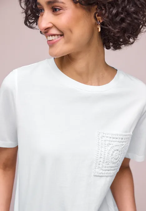 Frau mit lockigem Haar und verschwommenem Gesicht trägt weißes T-Shirt mit gehäkelter Tasche.