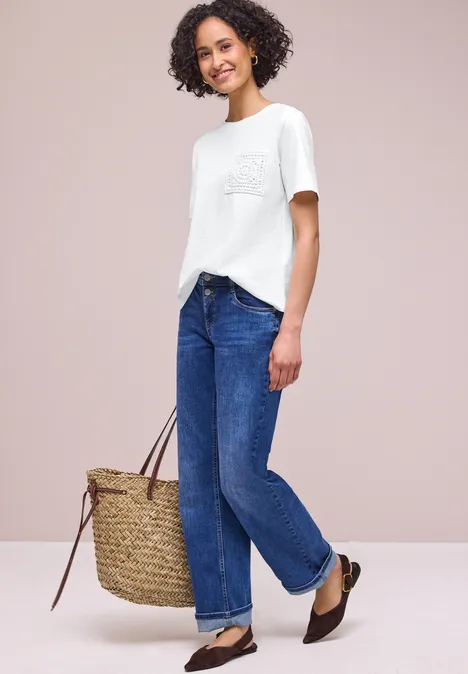 Frau in weißem T-Shirt, blauen Jeans und braunen Ballerinas mit großer Korbtasche.