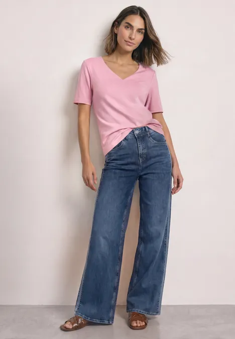 Kurzarm Shirt mit Herzausschnitt vintage blush Frau in rosa V-Shirt und weiten Jeans steht vor neutralem Hintergrund.