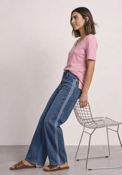 Kurzarm Shirt mit Herzausschnitt vintage blush Frau in rosa T-Shirt und zweifarbigen Jeans lehnt an modernem Drahtstuhl vor weißem Hintergrund.
