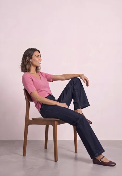 T-shirt manches courtes à col cœur warm blush Frau sitzt entspannt seitlich auf einem Holzstuhl, trägt rosa Shirt, blaue Jeans und braune Schuhe vor hellem Hintergrund.