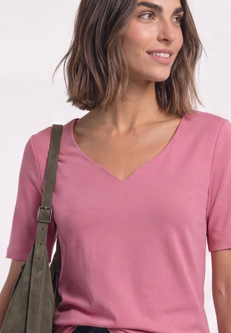 T-shirt manches courtes à col cœur warm blush Frau in rosa V-Ausschnitt-Shirt trägt grüne Schultertasche und lächelt seitlich.