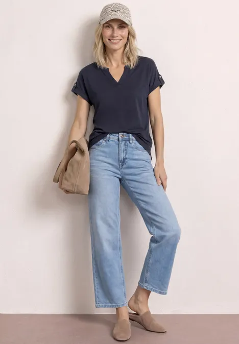 T-Shirt mit Split Neck und Knopfdetails shadowed navy Frau in weitem Jeans, schwarzem V-Neck Shirt, Beigetönen und Wildleder-Mules, hält Jacke über der Schulter