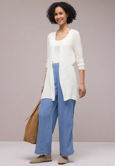 Een persoon draagt een lange witte cardigan, wijde blauwe broek, beige espadrilles en een bruine tas.