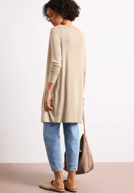 Achteraanzicht van een vrouw in een beige lange cardigan, blauwe jeans en instappers, met een beige tas