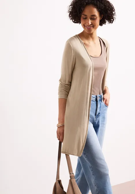 Vrouw in een lange beige cardigan, een bruin topje, lichte spijkerbroek en een schoudertas.