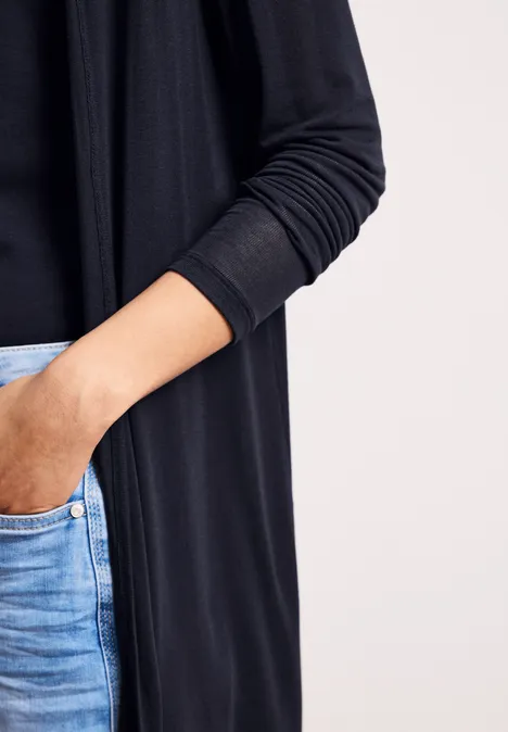 Iemand draagt een zwarte cardigan en een blauwe jeans, met de rechterhand nonchalant in de voorzak.