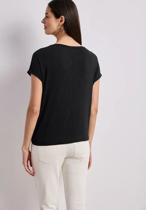 Frau von hinten in schwarzem T-Shirt und weißen Hosen vor hellem Hintergrund.