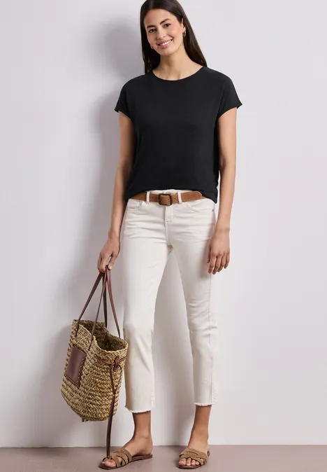 Frau in schwarzem Shirt, weißen Hosen, flachen Sandalen und Flecht-Tasche vor weißer Wand.