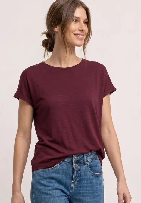 Een vrouw draagt een wijnrood T-shirt en blauwe jeans voor een lichte achtergrond.