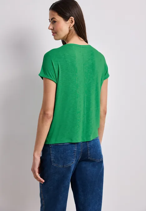 Frau trägt grünes T-Shirt und blaue Jeans, steht mit dem Rücken zur Kamera vor hellem Hintergrund.