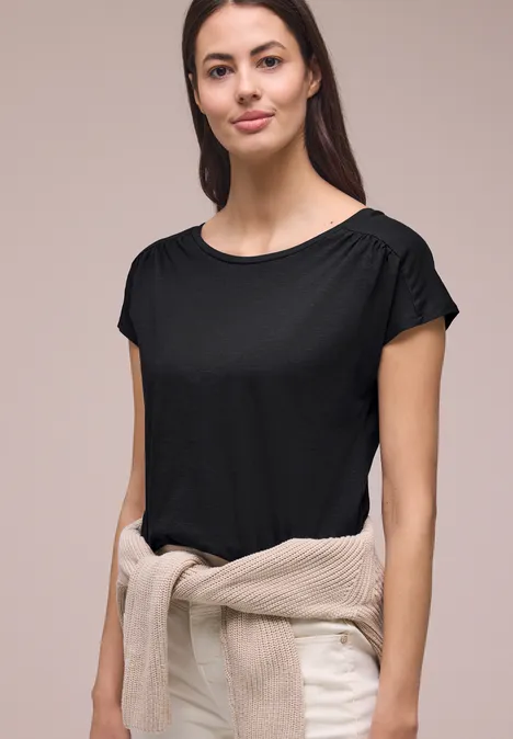 Shirt met ronde hals en rimpels Black Vrouw in zwart T-shirt met korte mouwen, beige trui om de taille geknoopt, neutrale achtergrond.