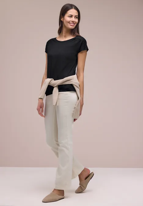 Shirt met ronde hals en rimpels Black Vrouw in zwart T-shirt, lichte broek, trui om de taille geknoopt en sandalen.