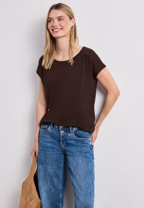 Eine junge Frau trägt ein schlichtes braunes T-Shirt, blaue Jeans und hält eine Tasche in der Hand.