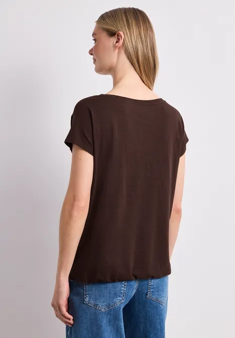 Frau von hinten in braunem T-Shirt und blauen Jeans vor hellem Hintergrund.