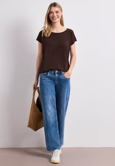 Frau in braunem T-Shirt, weitem Jeans, weißen Sneakern, mit brauner Tasche vor hellem Hintergrund.