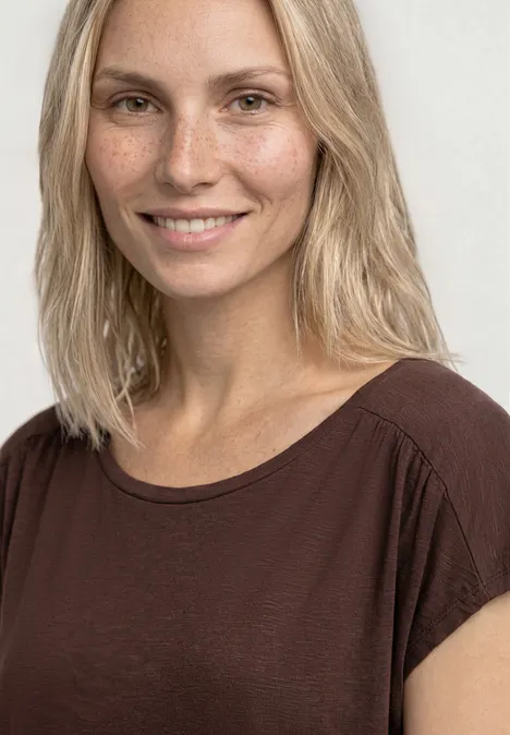 Eine blonde Frau trägt ein braunes T-Shirt vor hellem Hintergrund.