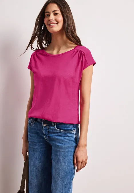 Vrouw draagt roze T-shirt en blauwe spijkerbroek voor een effen lichte achtergrond.
