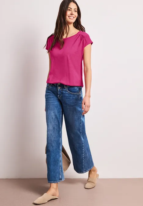 Vrouw in roze t-shirt en wijdvallende jeans poseert voor een lichte achtergrond.