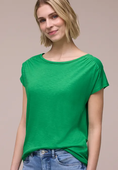 Eine Person trägt vor hellem Hintergrund ein grünes T-Shirt und Jeans.